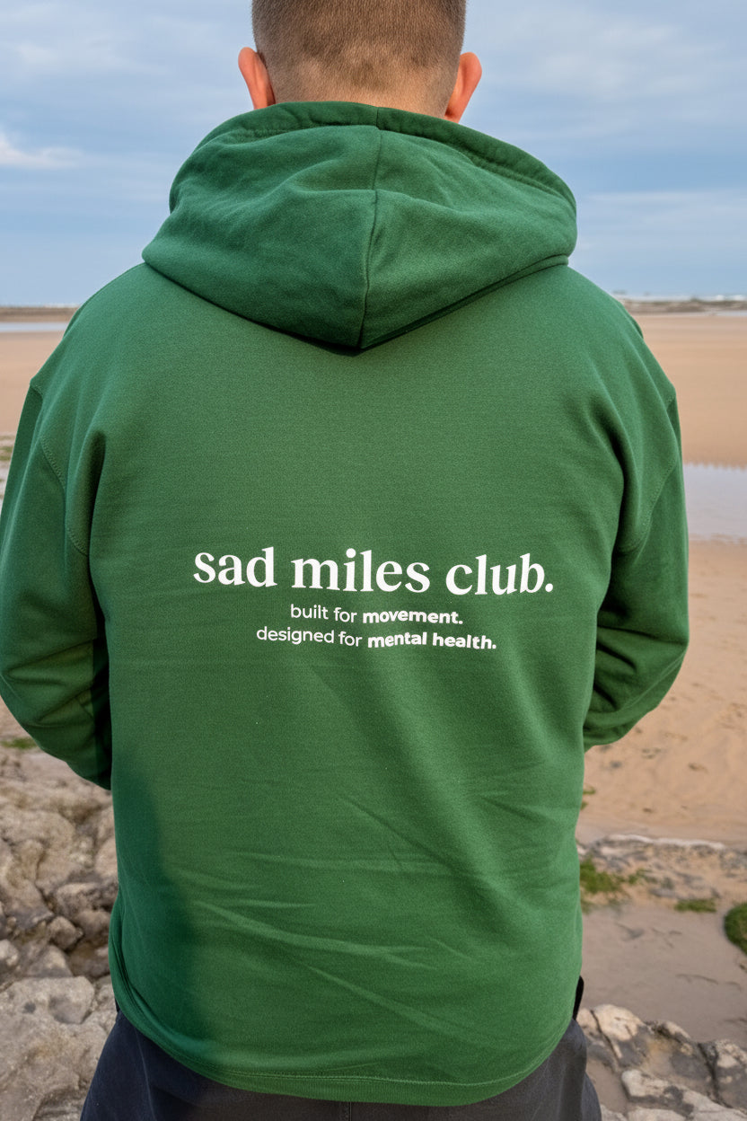 Person wearing a green hoodie with 'sad miles club' text on the back, standing on a beach.