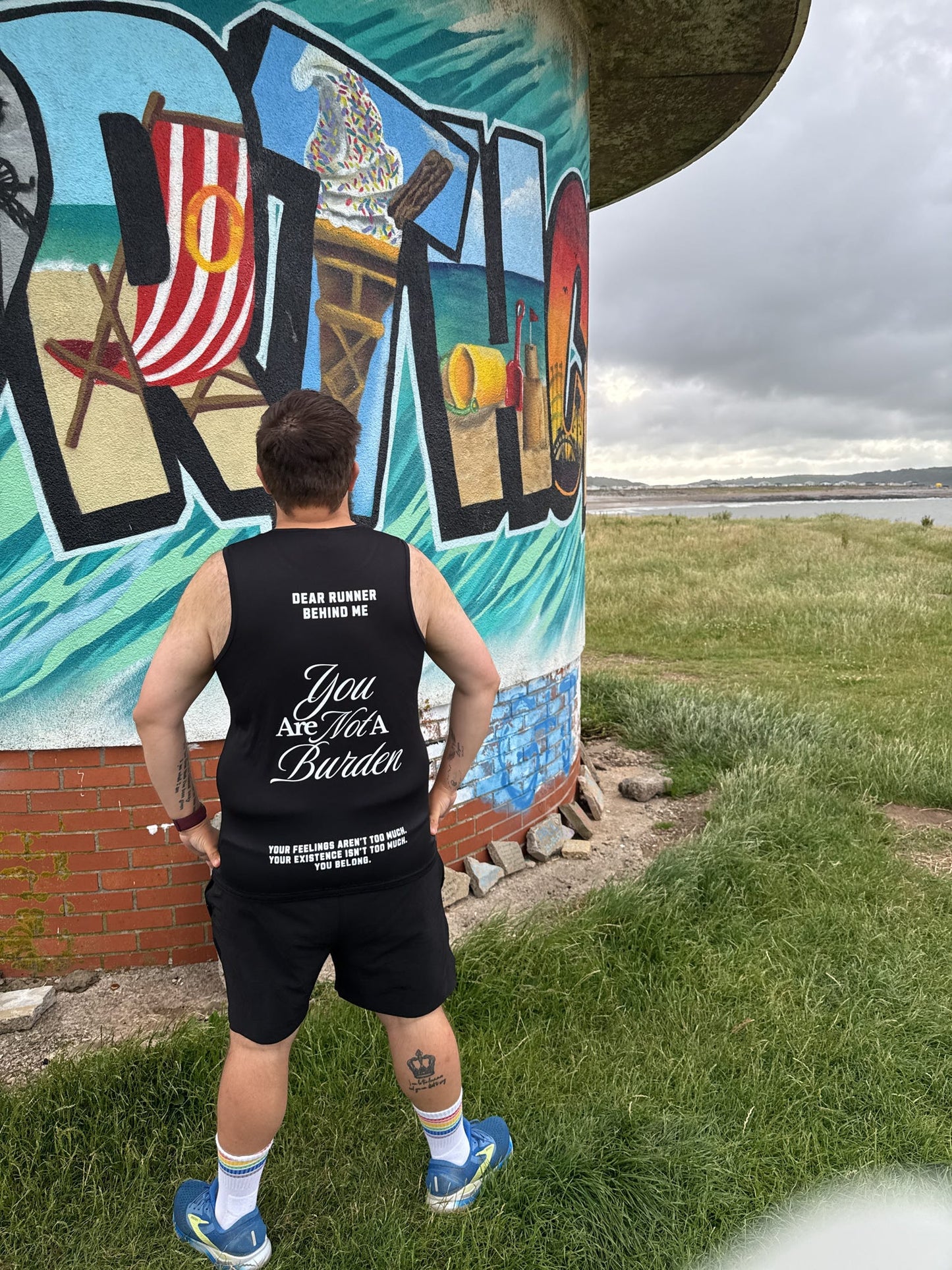 Person wearing a black tank top with text standing in front of a colorful mural.