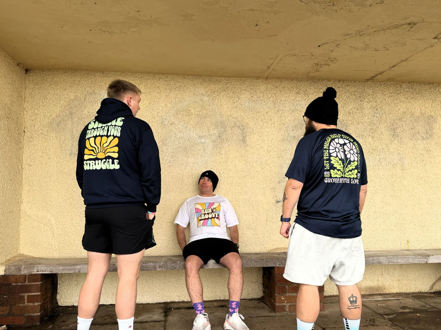 Three people in running gear with graphic tees and hoodies, motivational text on clothing, under shelter.