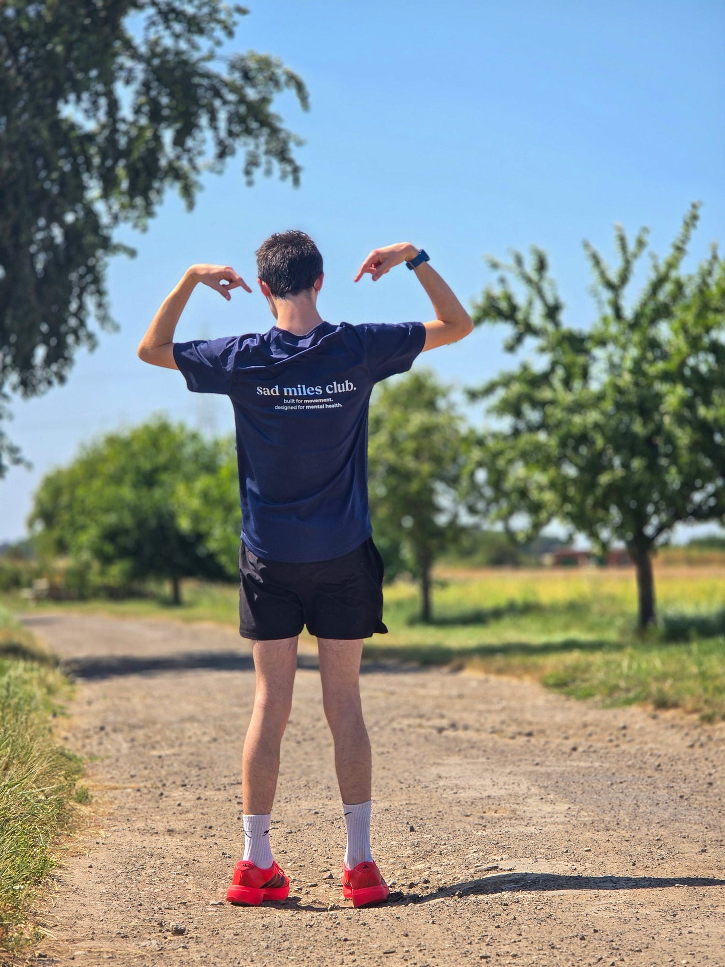 A man standing on a dirt path outdoors, viewed from behind, wearing a navy blue t-shirt with white text that reads 'bad milles club', black shorts, white socks, and red sneakers. Trees and a blue sky are visible in the background.