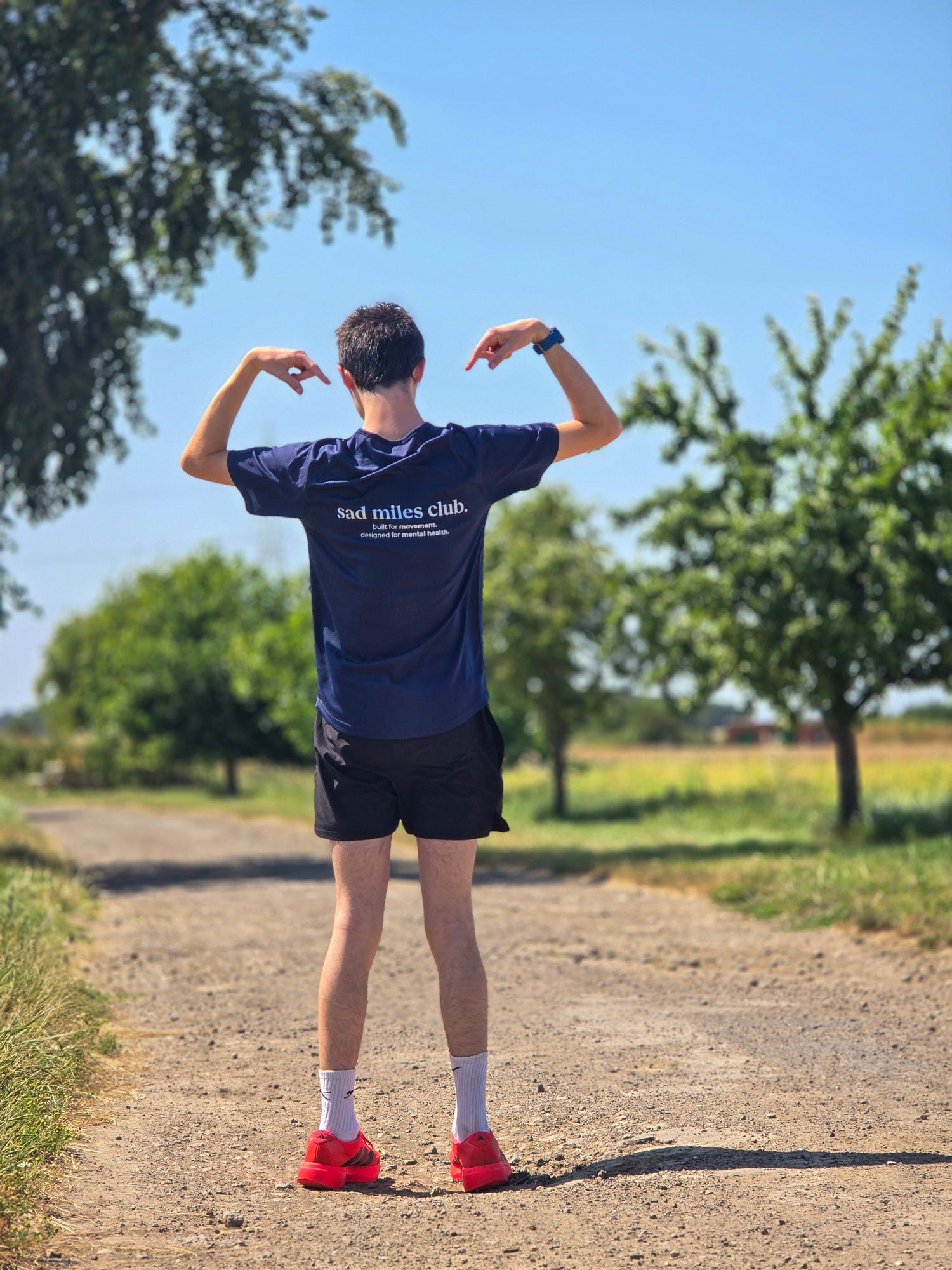 A man standing on a dirt path outdoors, viewed from behind, wearing a navy blue t-shirt with white text that reads 'bad milles club', black shorts, white socks, and red sneakers. Trees and a blue sky are visible in the background.