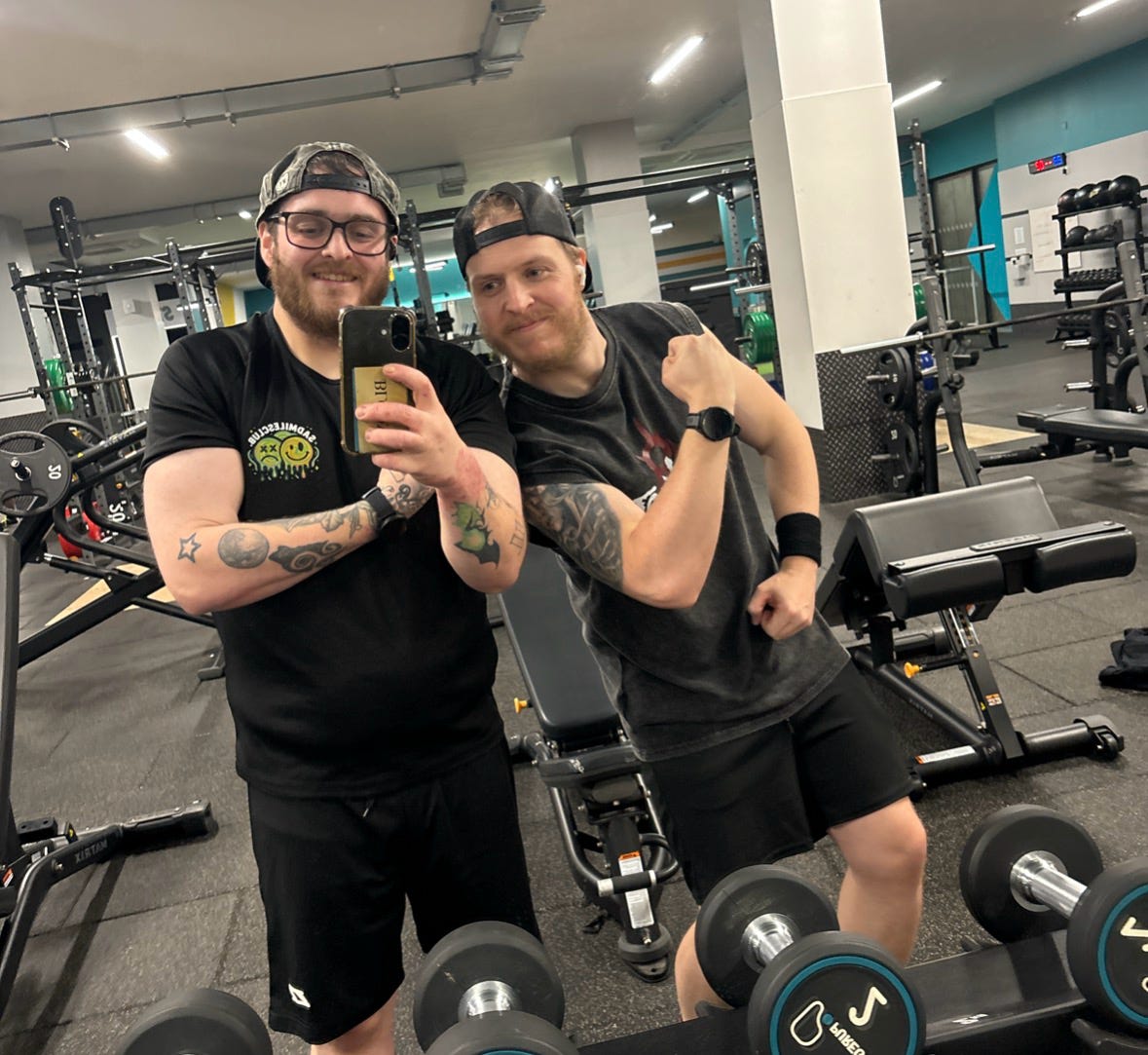 Two men taking a selfie in a gym with weights and exercise equipment around.