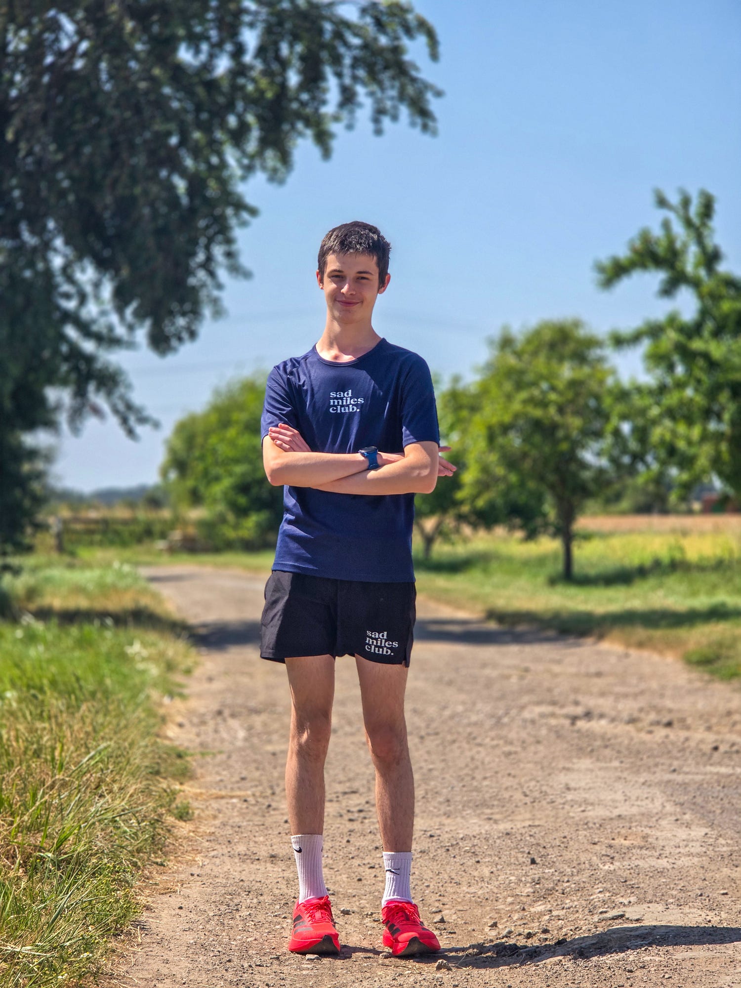 A man standing on a dirt path outdoors wearing a navy blue t-shirt with 'Tissot' printed on the chest, black athletic shorts, white socks, and bright red running shoes.
