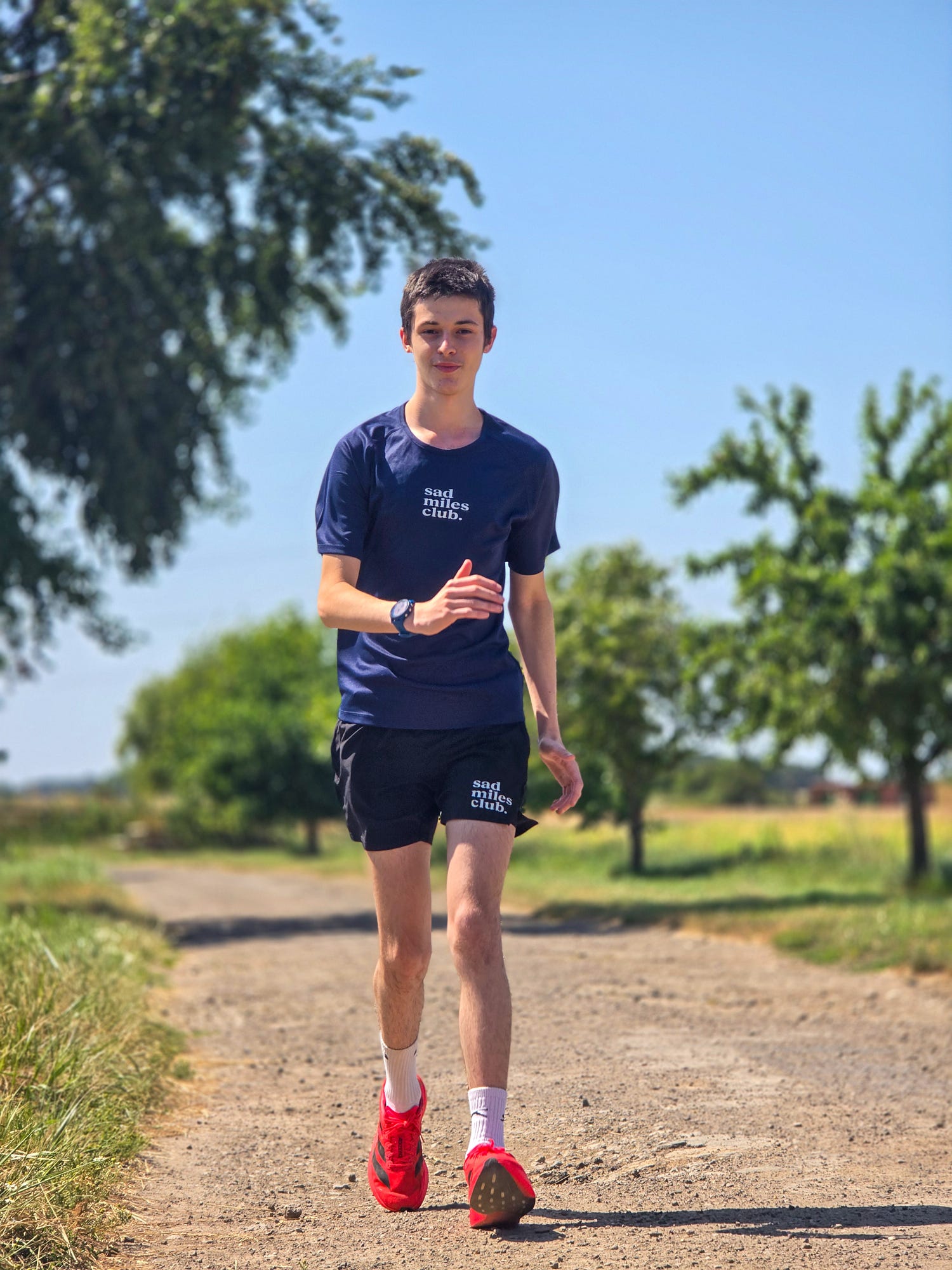A man running outdoors on a dirt path wearing a navy blue t-shirt with white text, black athletic shorts, white socks, and bright red running shoes.