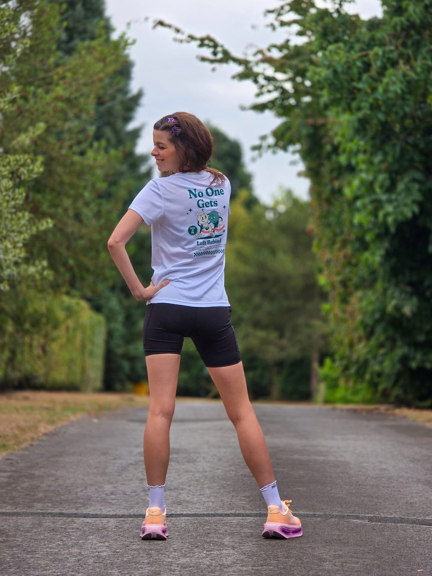 A woman standing on a paved path outdoors, viewed from behind, wearing a white graphic T-shirt with the text 'No One Gets Left Behind' and a cartoon illustration on the back, paired with black athletic shorts, white socks, and pink and yellow running shoes.