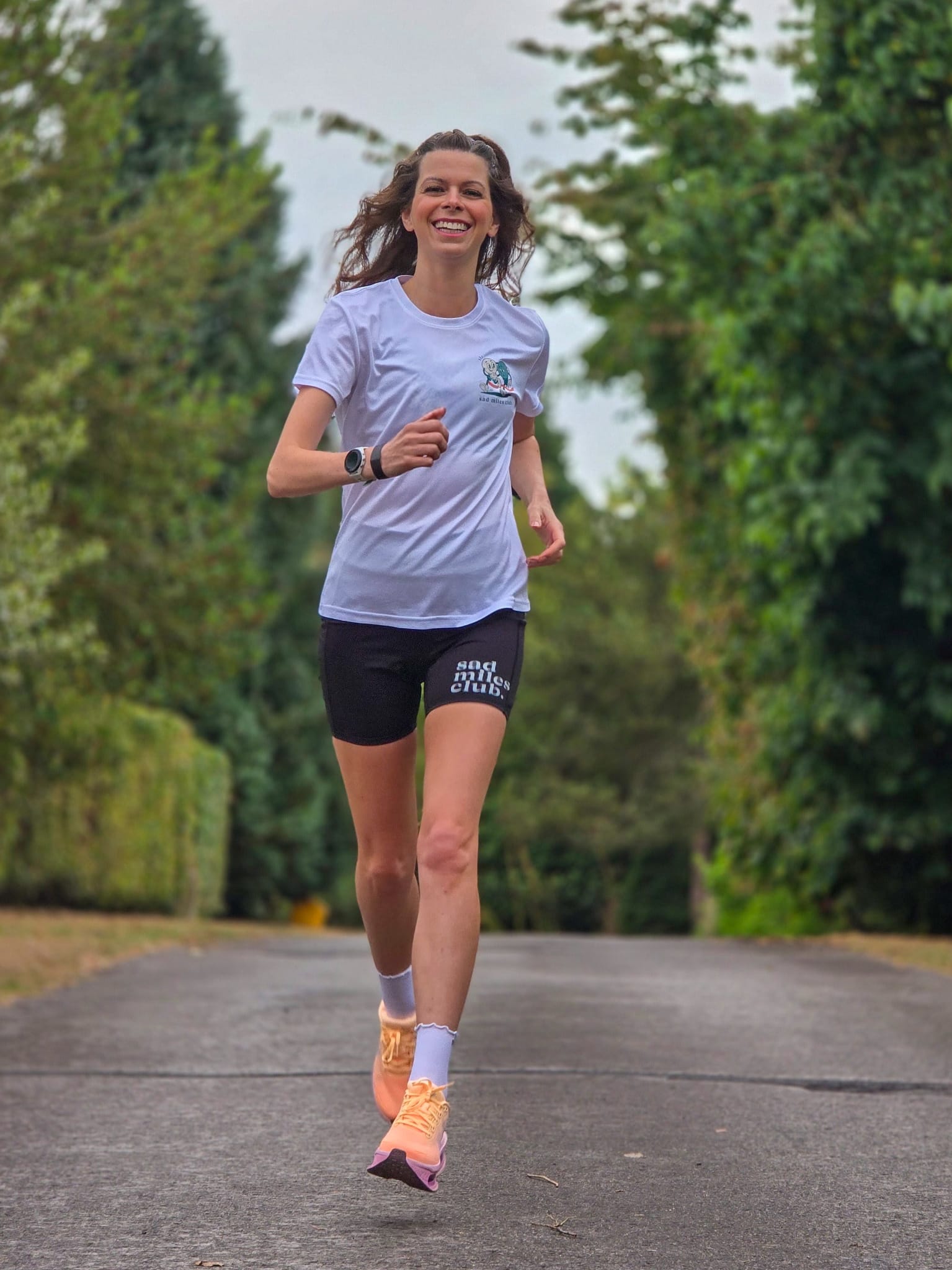 A woman running on an outdoor paved path wearing a light blue athletic t-shirt, black running shorts with white text, white socks, and orange running shoes.