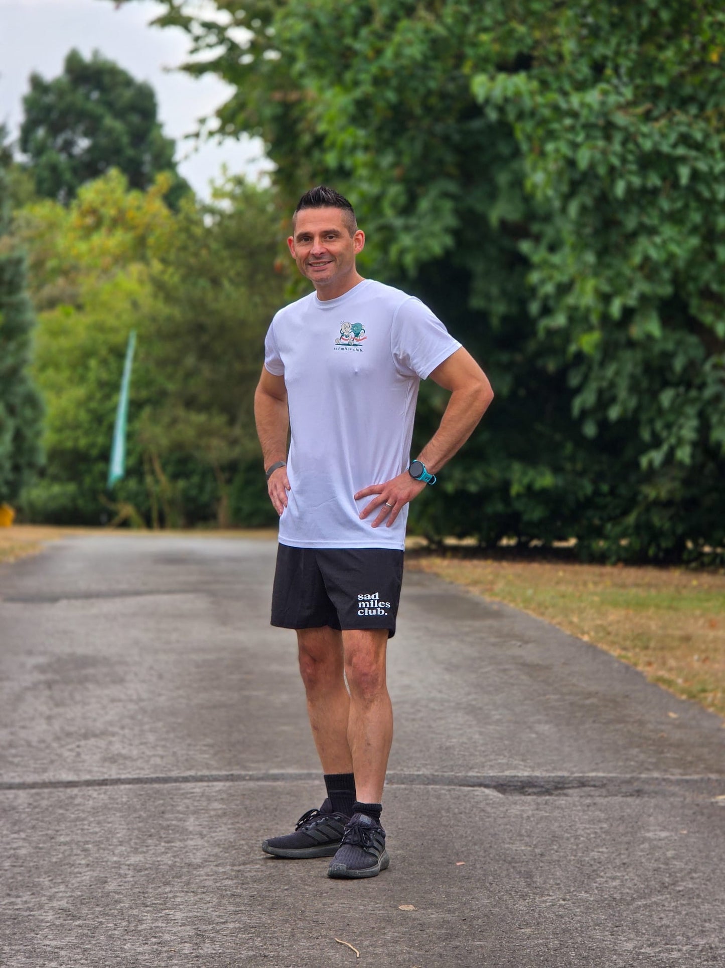 A man standing outdoors on a paved path wearing a white athletic t-shirt with a green and blue logo, black athletic shorts with white text, black socks, and black running shoes. The background shows green trees and grass.