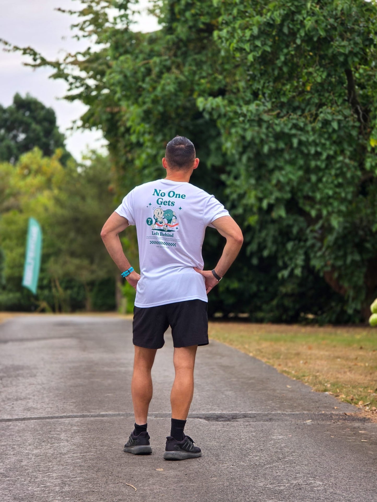 A man standing on an outdoor paved path with trees in the background, viewed from behind. He is wearing a white graphic T-shirt with blue and green text on the back, black athletic shorts, black socks, and black running shoes.