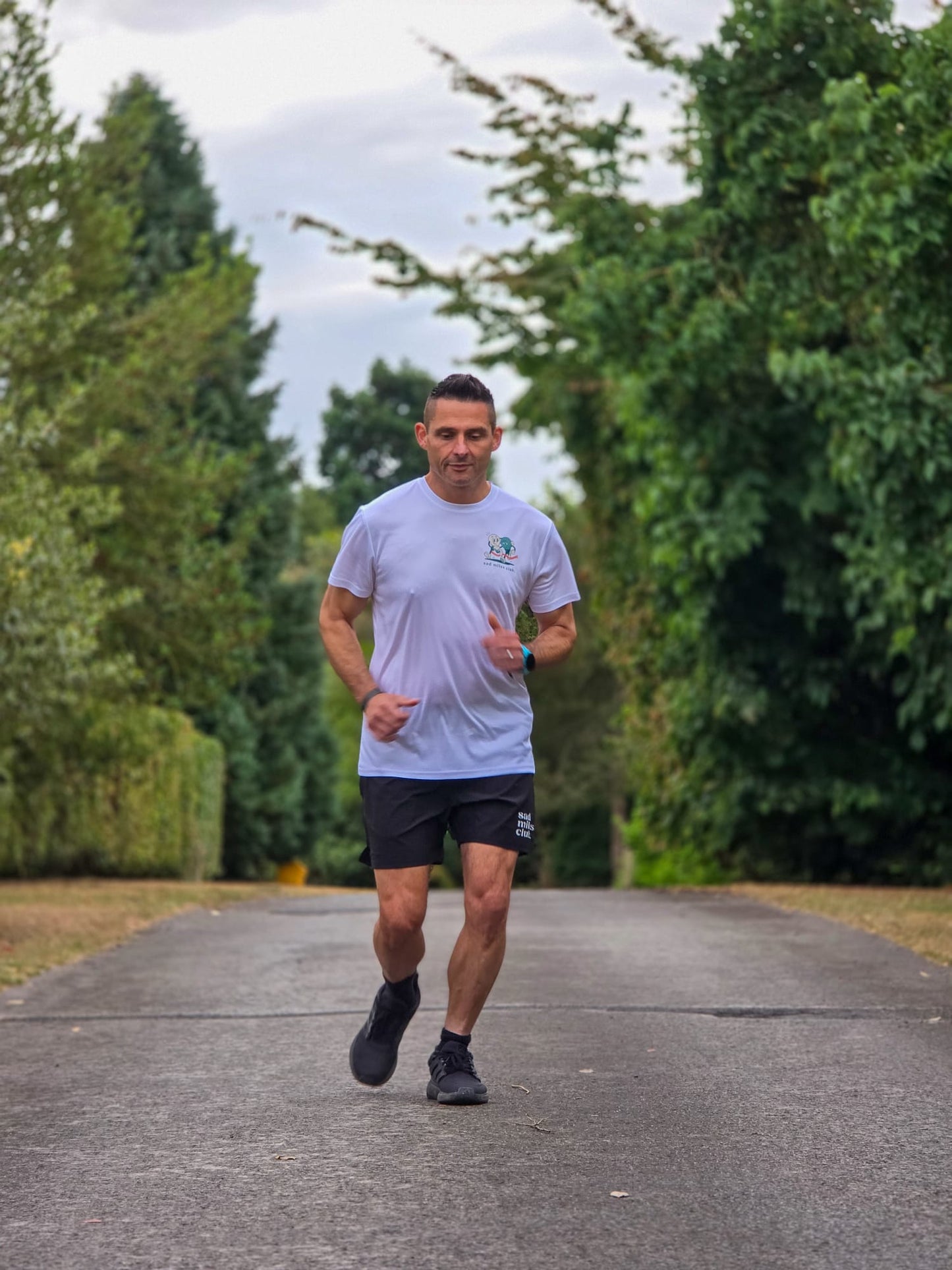 A man running on a paved path surrounded by green trees, wearing a light gray t-shirt, black athletic shorts, and black running shoes.