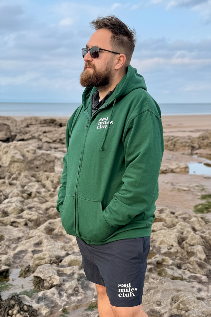 Man wearing a green hoodie and dark shorts with text, standing on a rocky beach.