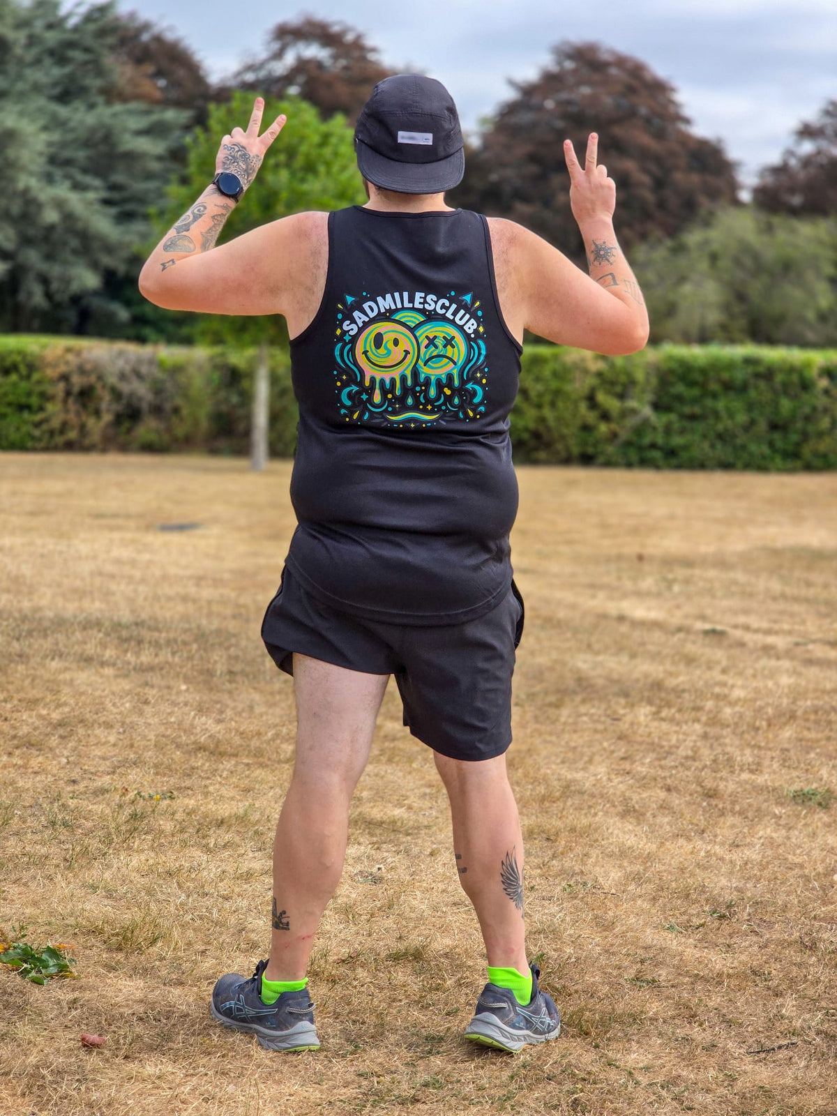 A person standing outdoors in a park wearing a black sleeveless tank top with a colorful 'Bad Mille Club' graphic on the back, black shorts, a black cap, and bright green socks.