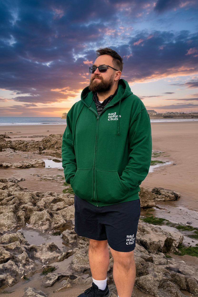 Man wearing a green hoodie and sunglasses standing on a rocky beach with a blue sky.