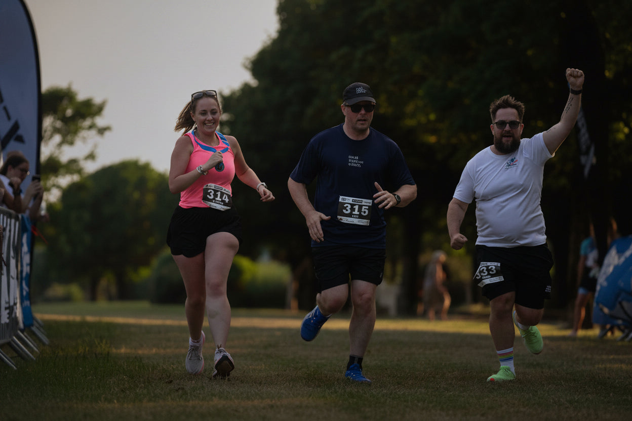 Three race participants running on a grassy course during an outdoor race, with trees and event banners in the background.