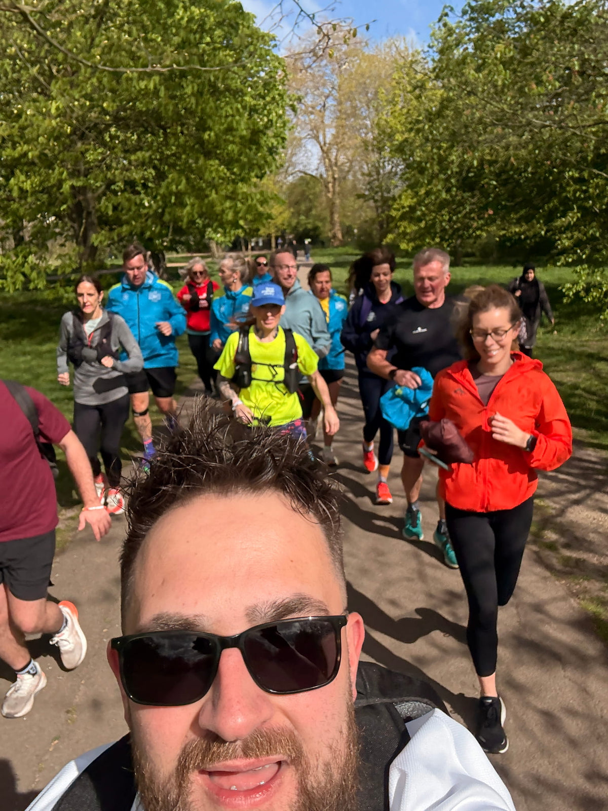 A group of runners jogging on a sunny park path, with a man in sunglasses taking a selfie in the foreground. The runners are wearing various athletic jackets and shirts in bright colors like blue, red, yellow, and black.