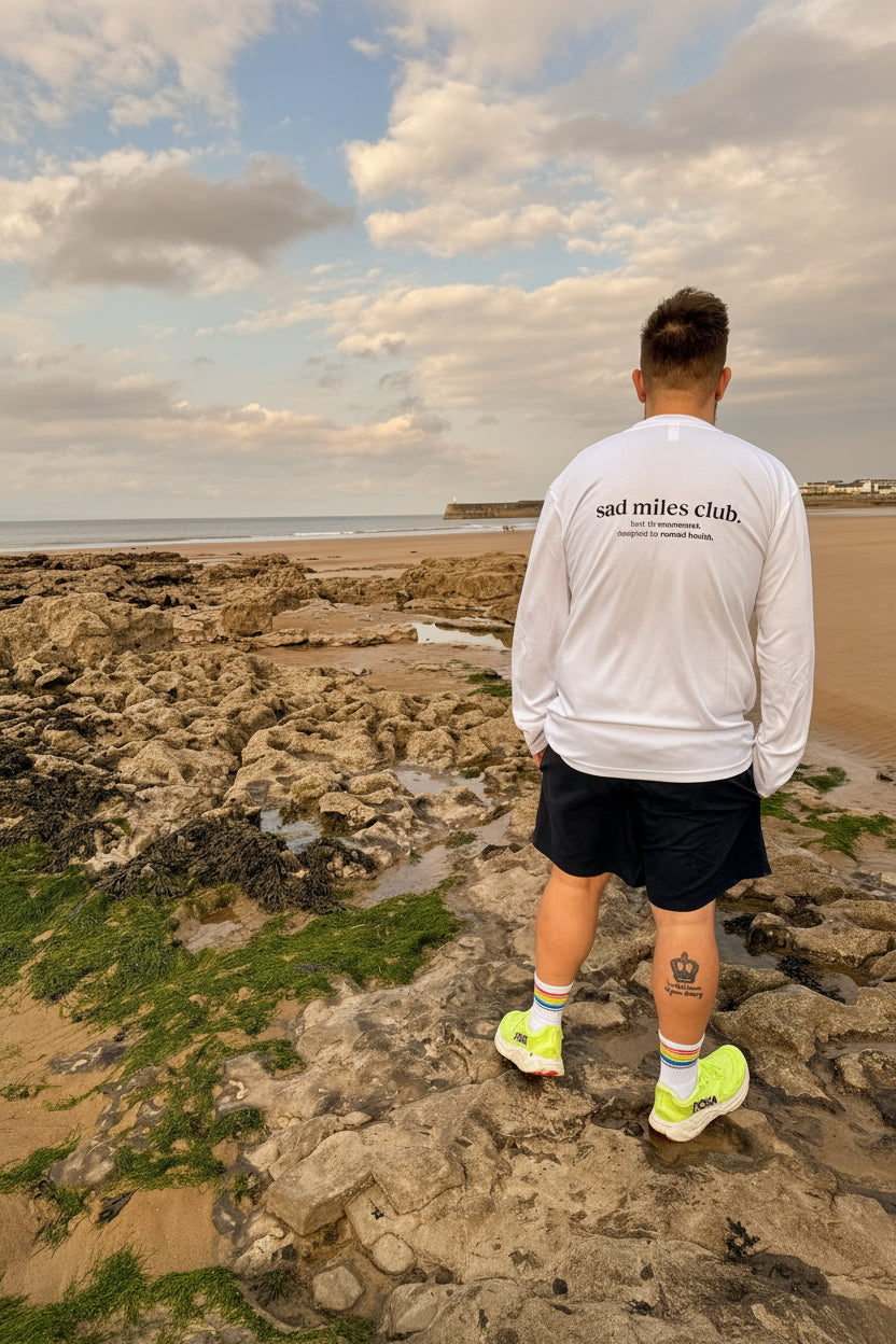 Person standing on a rocky beach wearing a white sweatshirt with text, shorts, and bright shoes.
