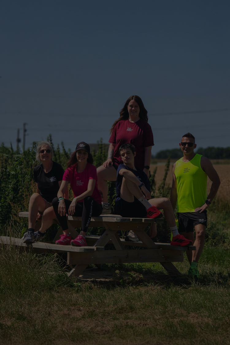 Five people in athletic wear posing together outdoors on a wooden picnic table in a grassy field under a clear blue sky.
