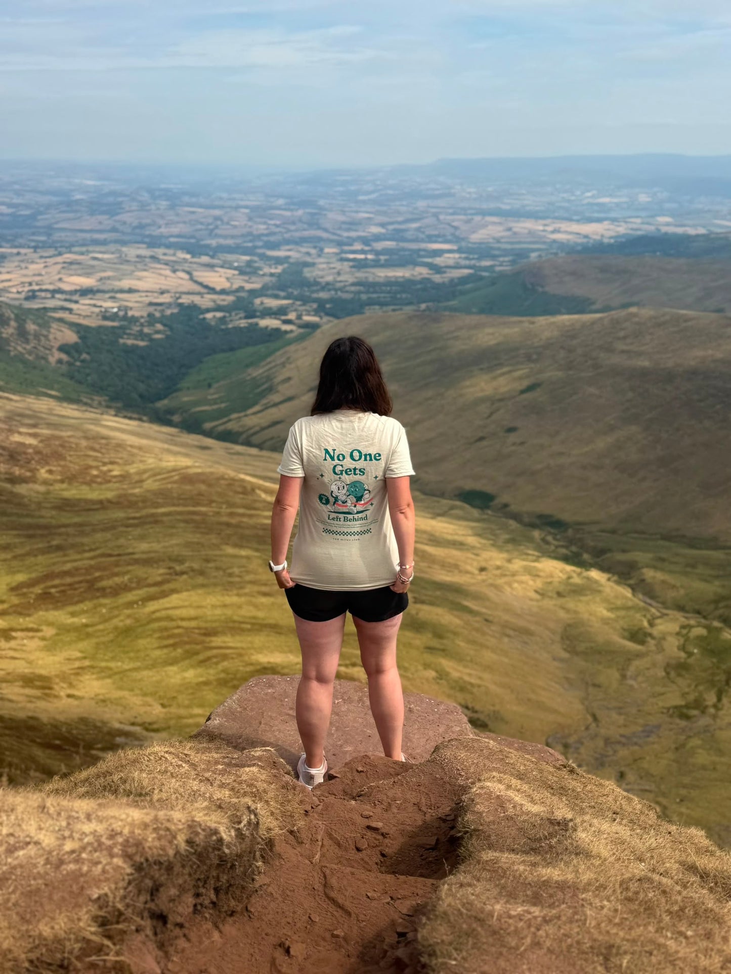A woman stands on a rocky cliff edge overlooking a vast, rolling green and brown valley landscape under a pale blue sky. She is seen from behind wearing a cream-colored graphic T-shirt with the text 'No One Gets' and a blue mountain illustration, paired with black shorts.