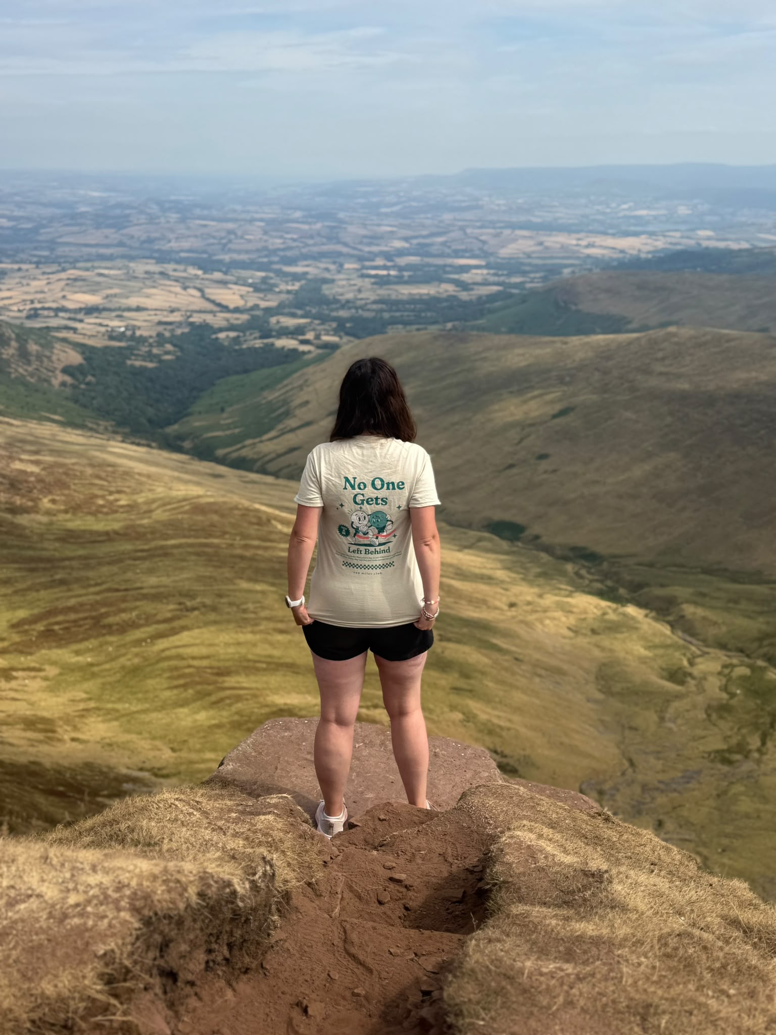 A woman stands on a rocky cliff edge overlooking a vast, rolling green and brown valley landscape under a pale blue sky. She is seen from behind wearing a cream-colored graphic T-shirt with the text 'No One Gets' and a blue mountain illustration, paired with black shorts.