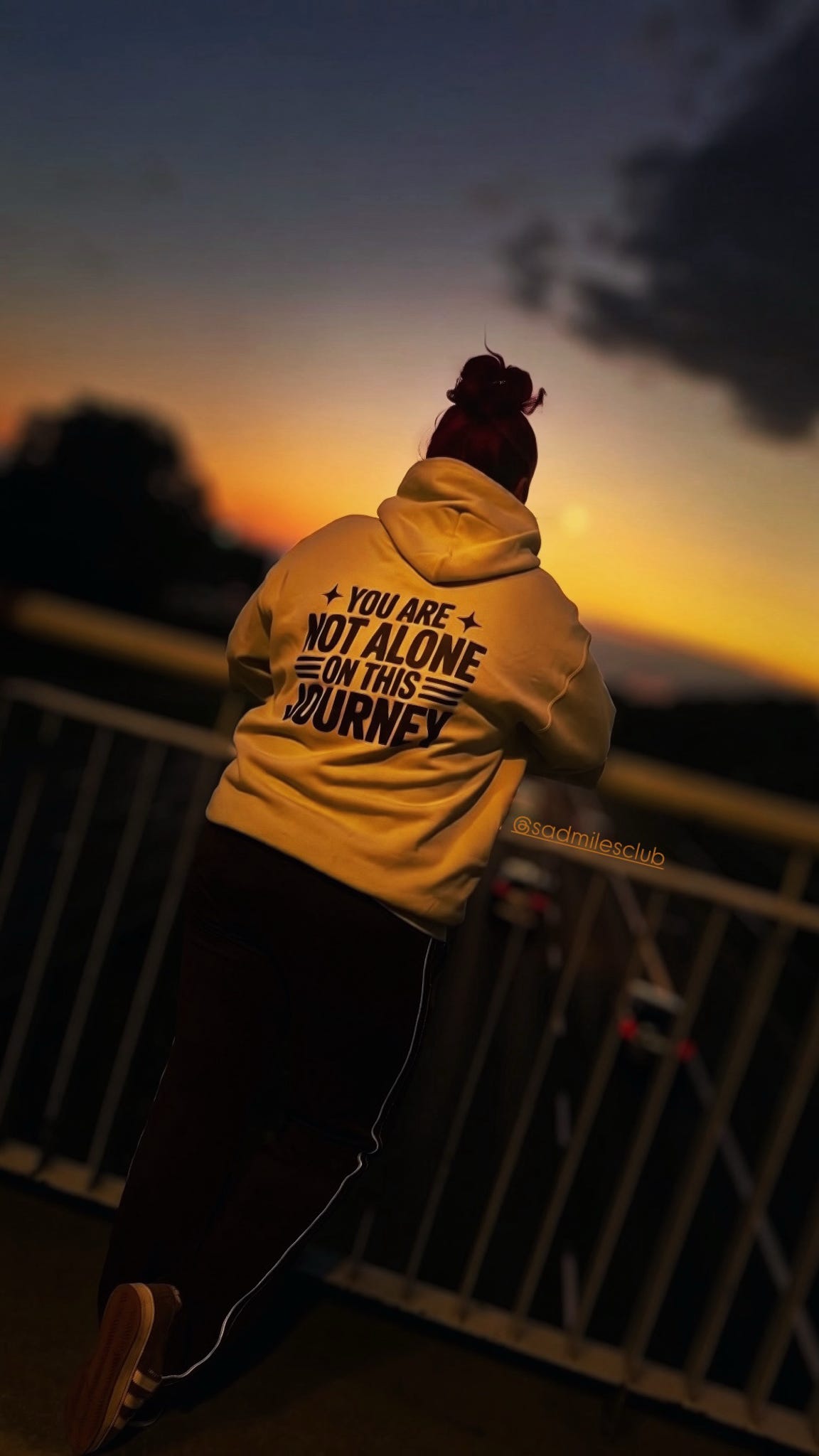 A person viewed from behind wearing a yellow hoodie with the text 'YOU ARE NOT ALONE ON THIS JOURNEY' printed in black, standing outdoors at sunset near a railing.