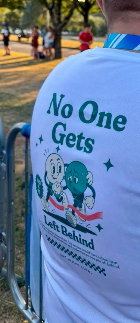 A man seen from behind wearing a white t-shirt with a blue and green graphic on the back that reads 'No One Gets Left Behind'. He is standing outdoors at sunset near a blue barrier, with trees and park activity in the background.