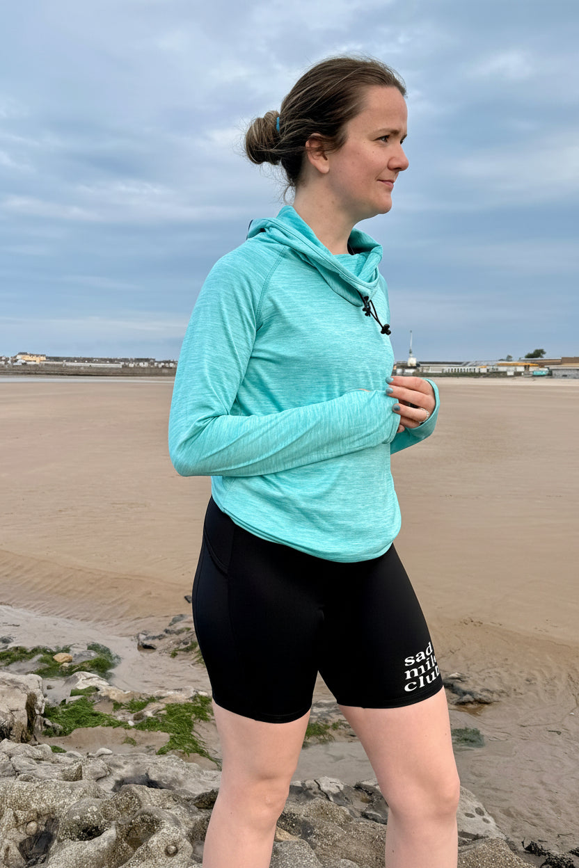 Woman in a light blue jacket and black shorts standing on a beach.