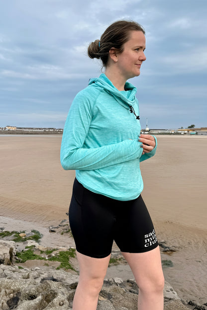 Woman in a light blue jacket and black shorts standing on a beach.