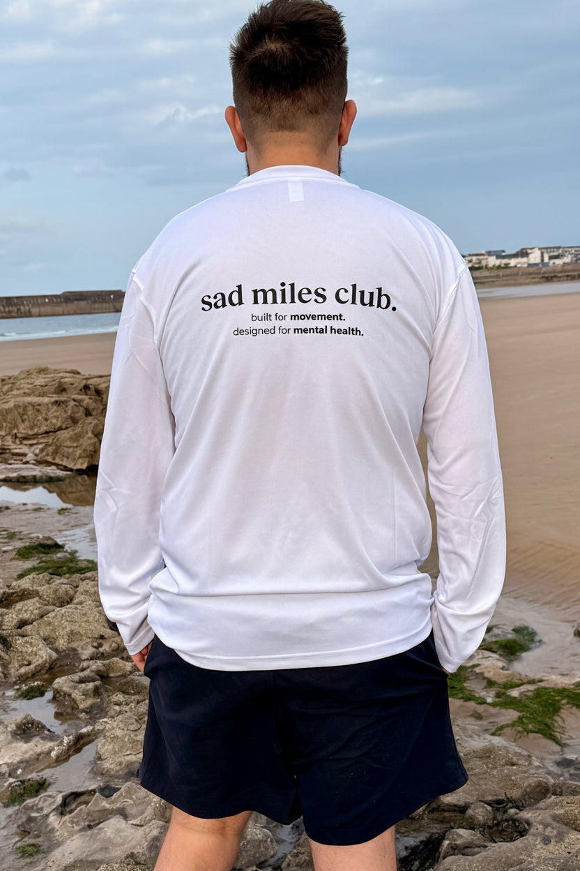 Person wearing a white long-sleeve shirt with 'sad miles club' text at a beach.