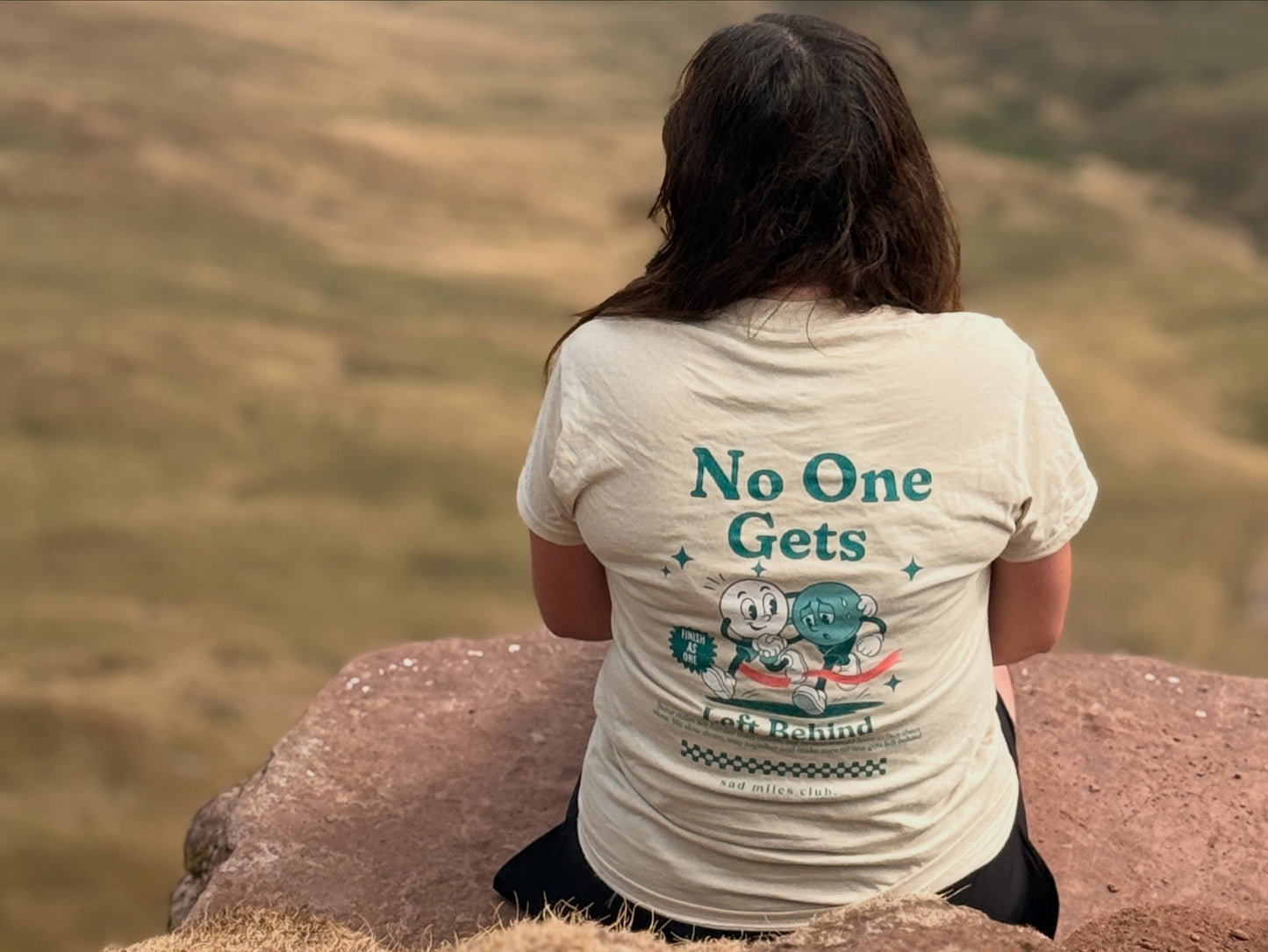 A person sitting on a rock outdoors, viewed from behind, wearing a cream-colored graphic t-shirt with the text 'No One Gets' and a cartoon character design on the back.