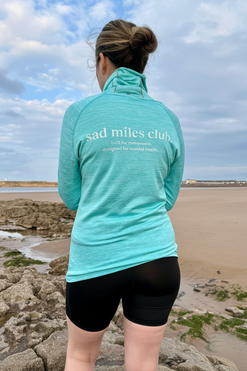 Person wearing a teal 'sad miles club' jacket at a beach.