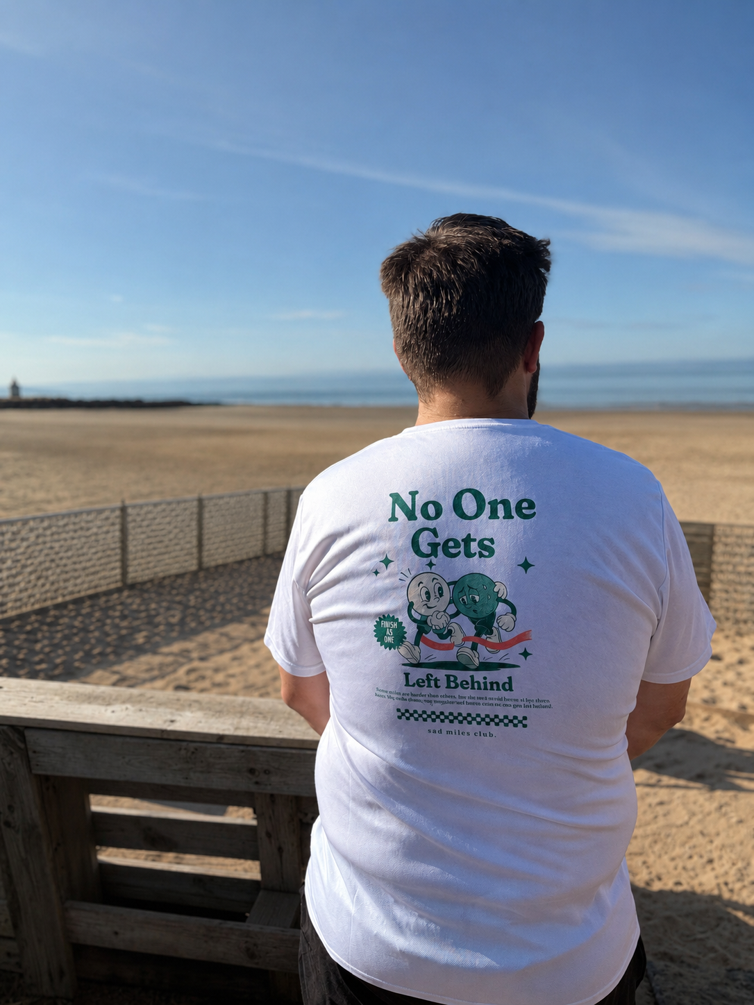 A man viewed from behind wearing a white graphic t-shirt with the text 'No One Gets Left Behind' and a cartoon character on the back, standing on a wooden boardwalk overlooking a sandy beach under a blue sky.