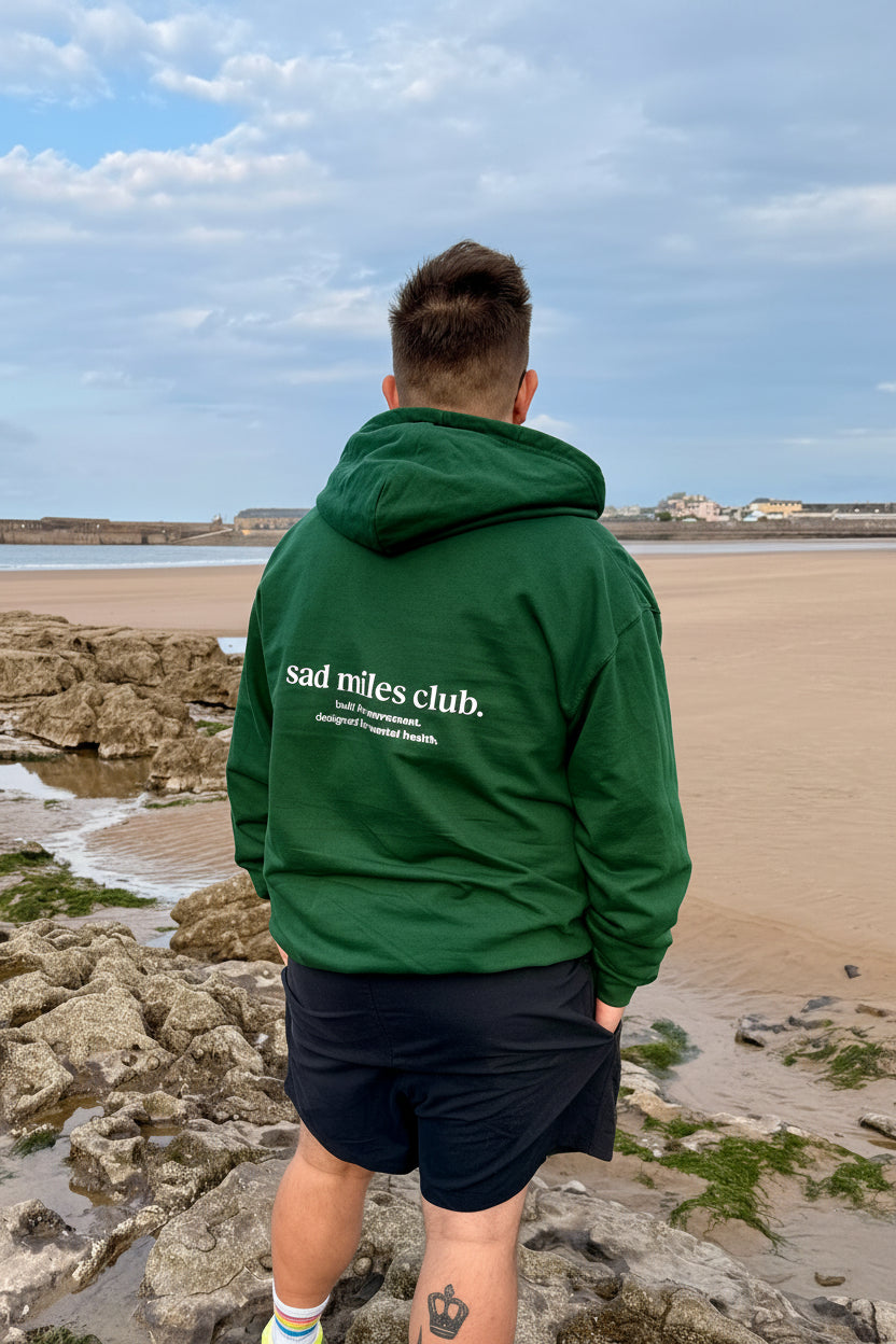 Person wearing a green hoodie with 'sad miles club' text on a beach