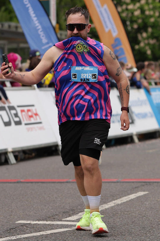 A man wearing a vibrant pink and blue wavy patterned sleeveless shirt, black shorts, and neon yellow running shoes walks across a white finish line at a marathon. The background shows a blurred street scene with spectators and banners.