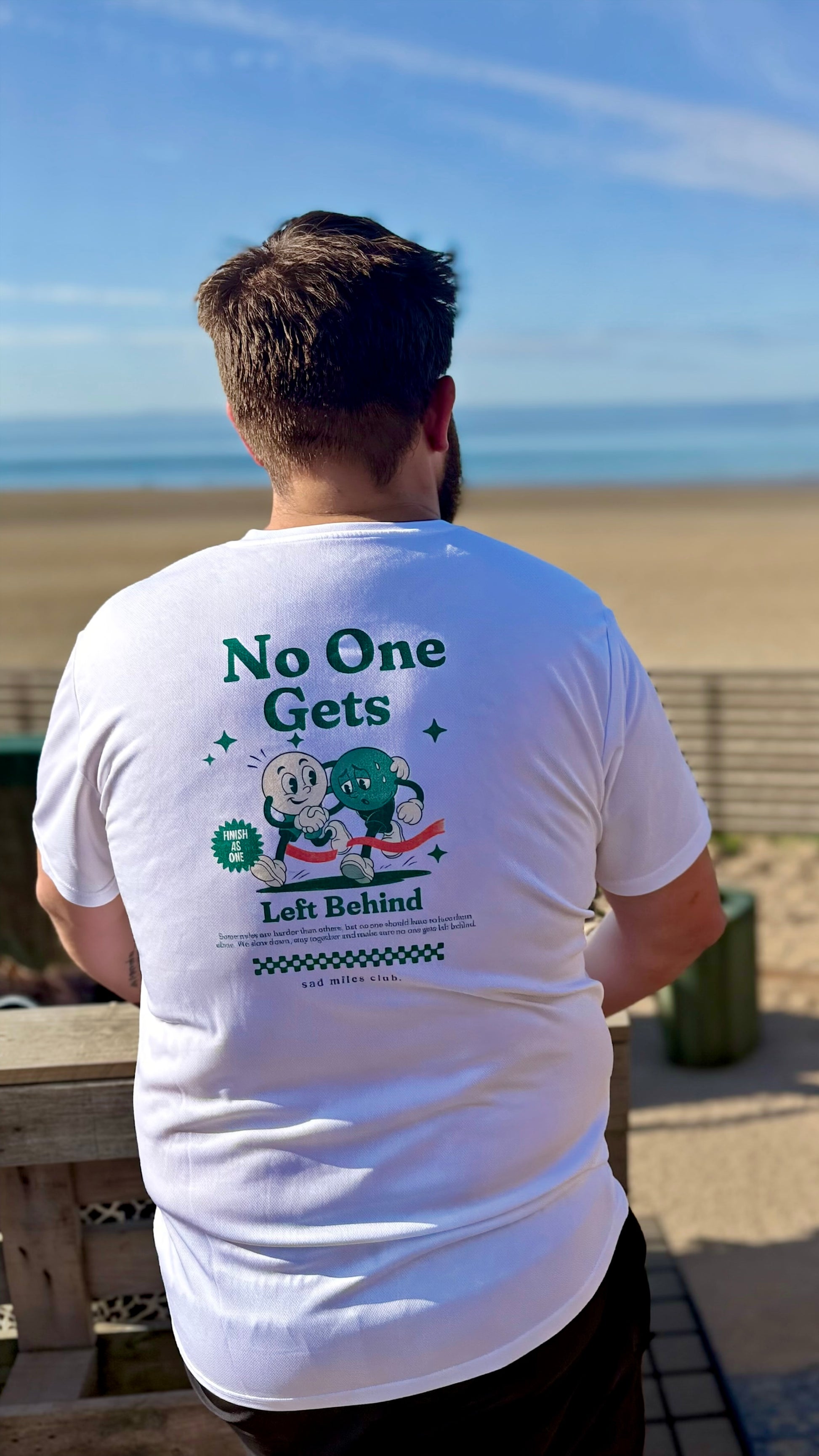 A man viewed from behind wearing a white graphic t-shirt with a green cartoon design and the text 'No One Gets Left Behind' on a beach background.