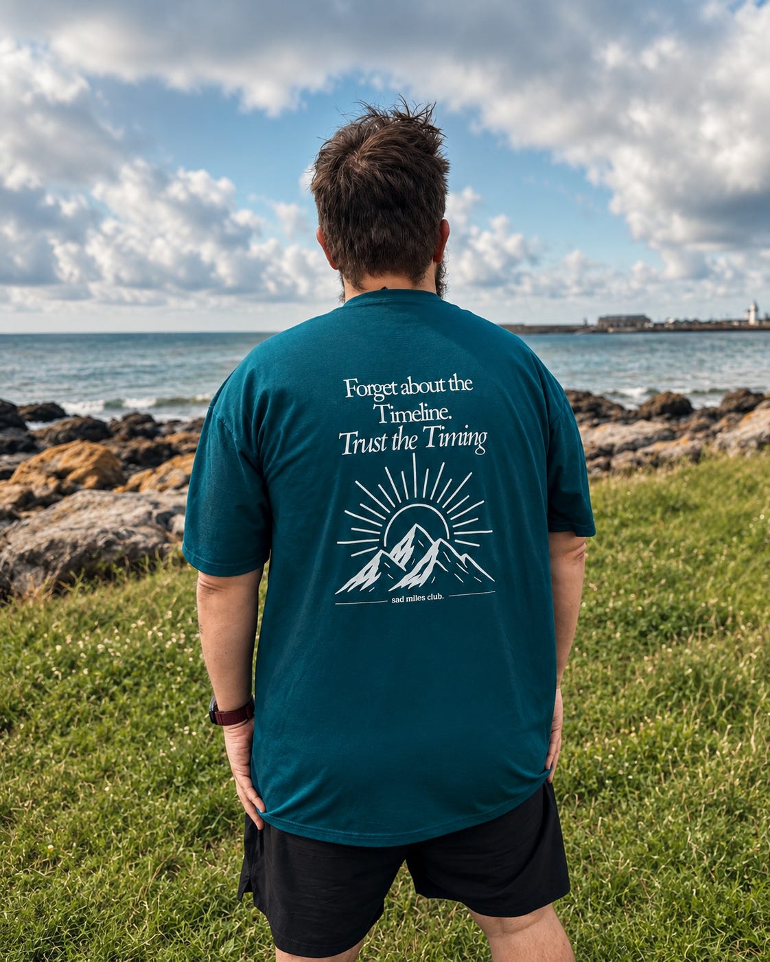 A man viewed from behind wearing a teal t-shirt with a white mountain graphic and the text 'Forget about the Timeline. Trust the Timing' on the back, standing on a grassy shore with the ocean and cloudy sky in the background.