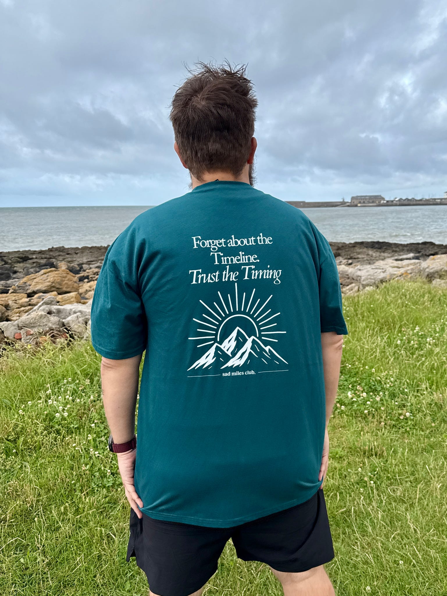 Person wearing a teal t-shirt with a motivational quote and mountain graphic, standing outdoors by a body of water.