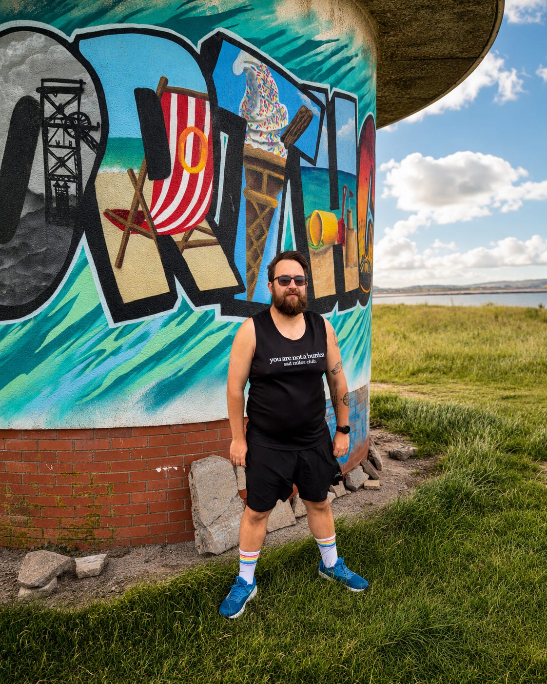 A bearded man wearing a black tank top, black shorts, blue sneakers, and white socks stands in front of a colorful graffiti-covered cylindrical structure outdoors on a grassy field under a blue sky.