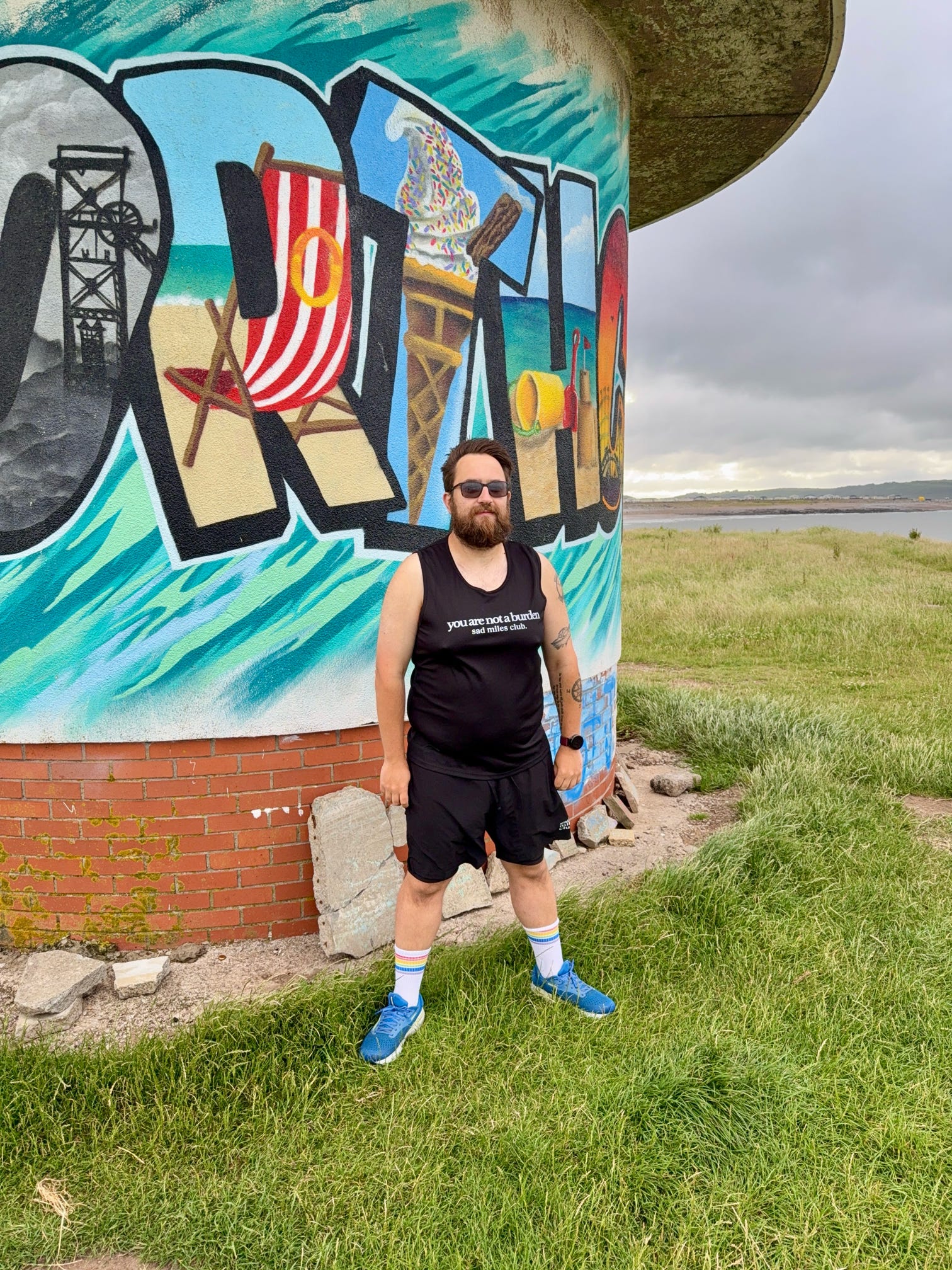 Man standing in front of a colorful mural with a scenic design on a brick wall.