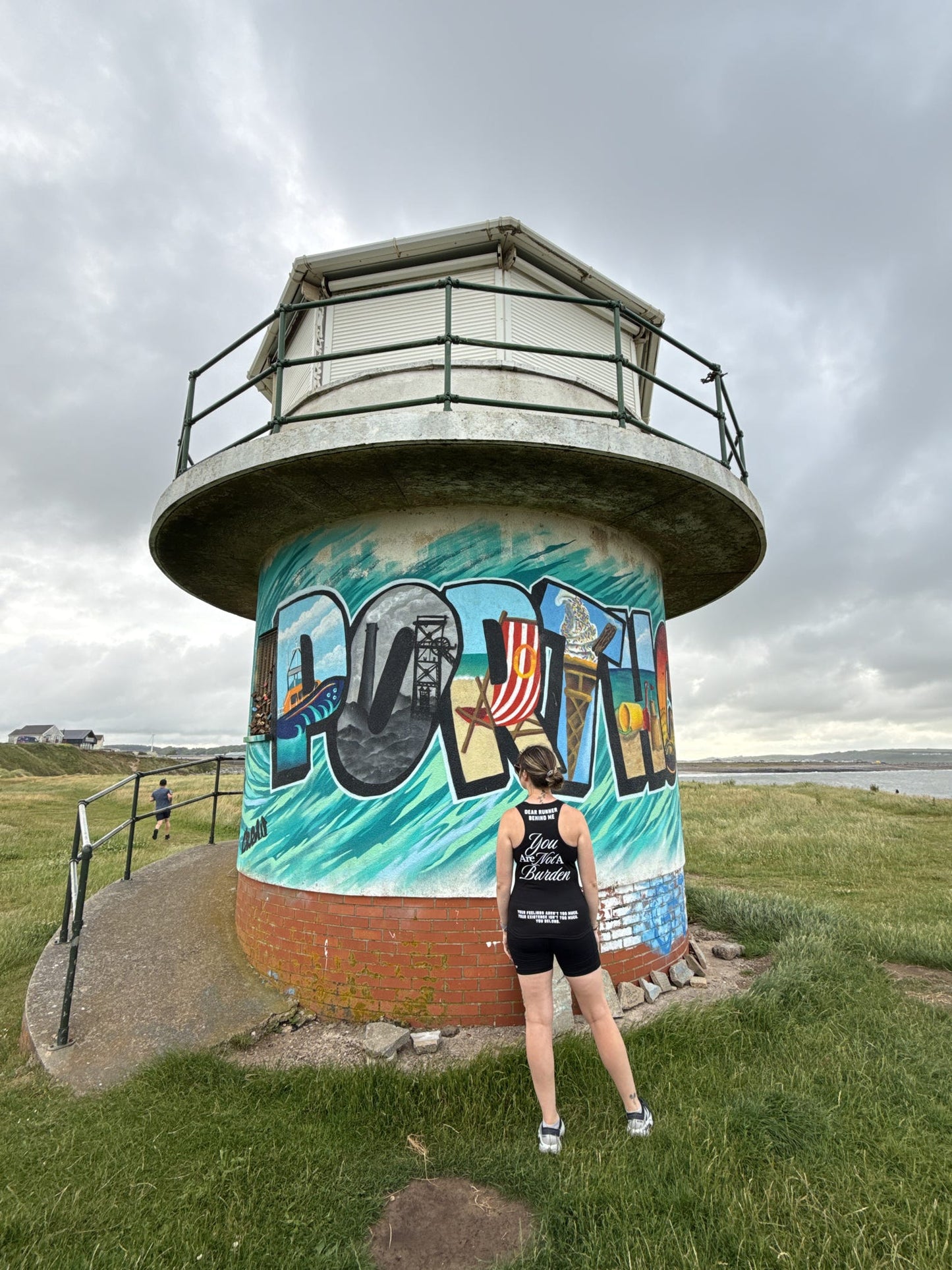 Person standing in front of a lighthouse with a colorful mural on a cloudy day.
