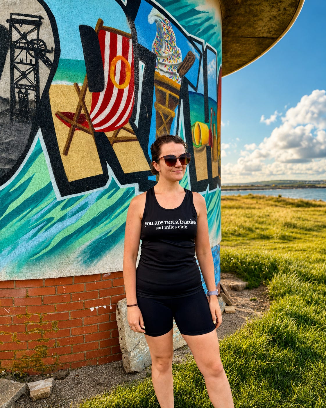 A woman wearing a black tank top with white text and black shorts stands in front of a colorful beach-themed mural outdoors next to a grassy field and water.