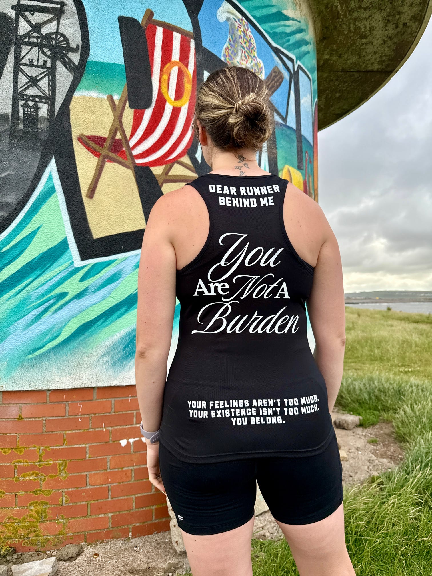 Person wearing a black tank top with motivational text in front of a colorful mural.