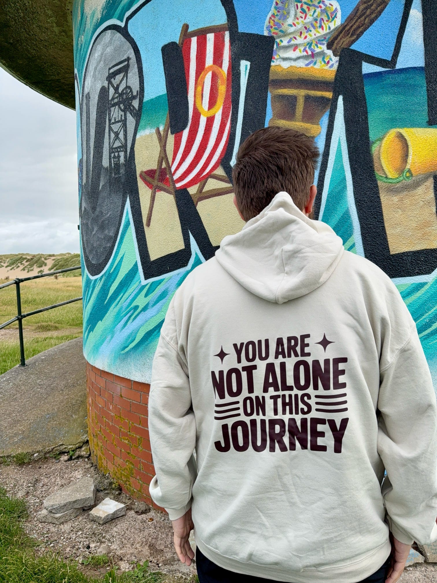 Person wearing a hoodie with 'You are not alone on this journey' text in front of a colorful mural.
